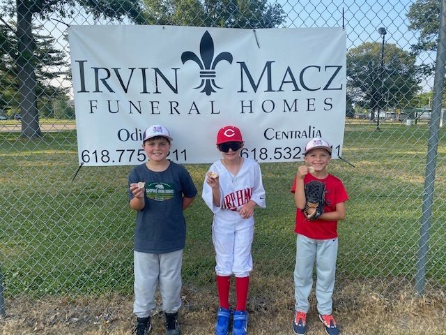 Three children in baseball uniforms in front of a funeral home banner, holding balls.