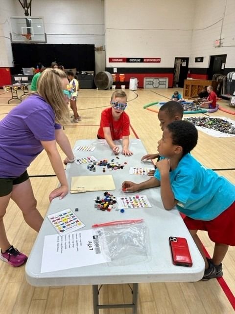 Children and adult at a table with Legos. Person in purple shirt watches three kids as they build. Indoor gym setting.