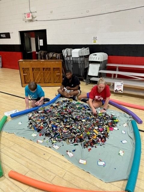 Three people seated around a pile of LEGOs inside a gym. Blue, orange, and green pool noodles form a circle.