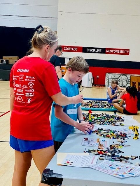 Woman in red shirt assisting a boy with LEGOs at a table in a gym. Other children in background.