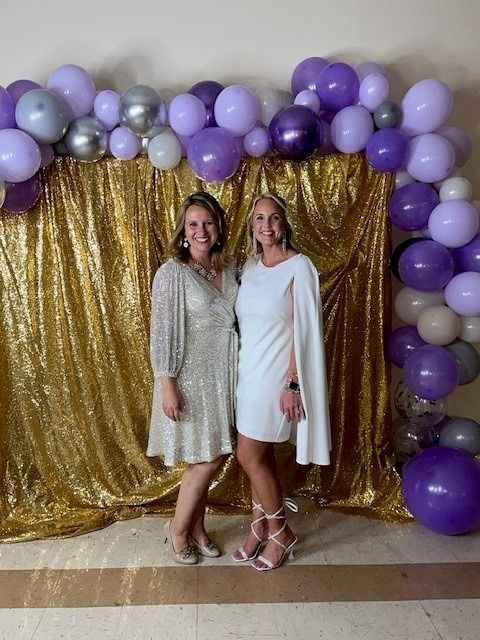 Two women pose in front of a gold sequin backdrop with purple and silver balloons. One wears a white dress, the other a silver dress.
