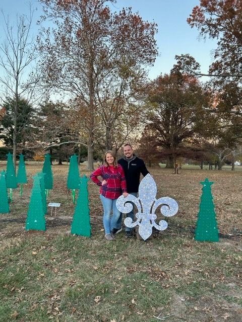 Couple posing with a large fleur-de-lis, surrounded by green Christmas tree cutouts in a grassy park setting.