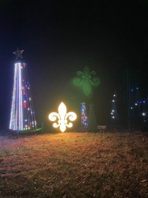 Illuminated holiday light display featuring a Christmas tree, fleur-de-lis, and other glowing shapes at night.