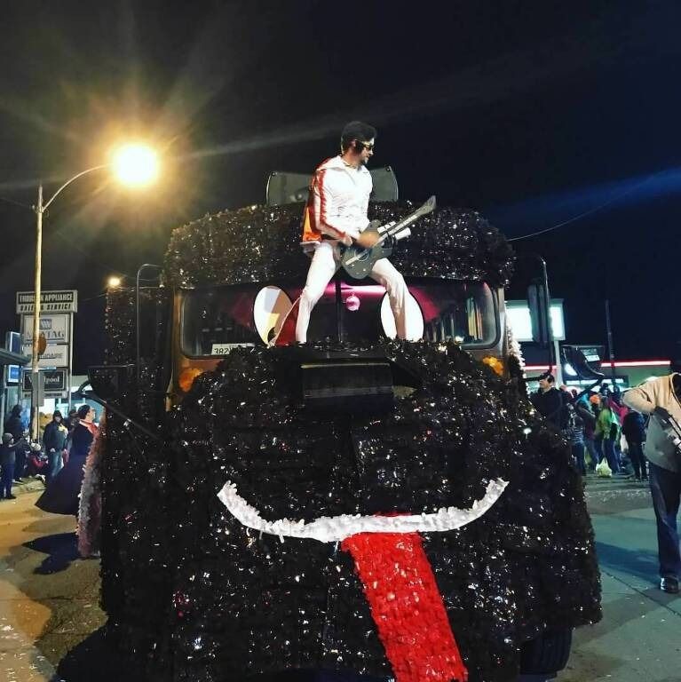 Man in Elvis-style outfit plays guitar atop a float decorated as a smiling, sequined black monster at night.