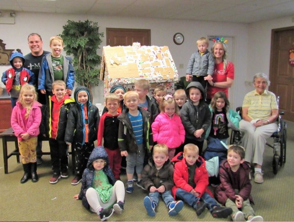 Children and adults pose with a gingerbread house. Indoors, children wear coats, smiles, and a woman sits in a wheelchair.