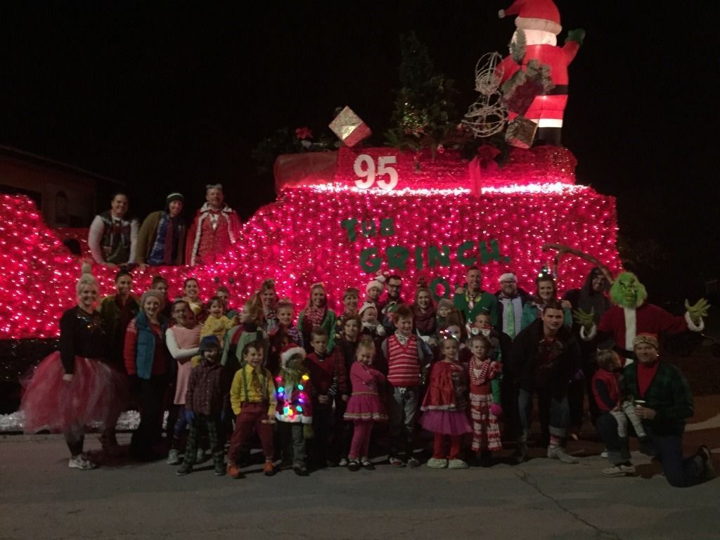 Group poses in front of a brightly lit Christmas float. Santa and the Grinch are present.
