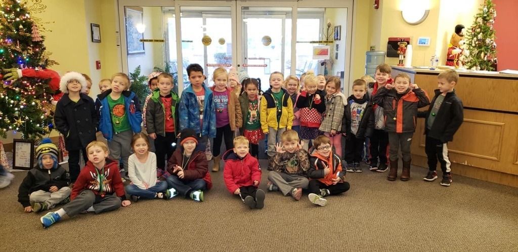 A group of children posing in front of glass doors, some sitting on the floor, near a decorated Christmas tree.