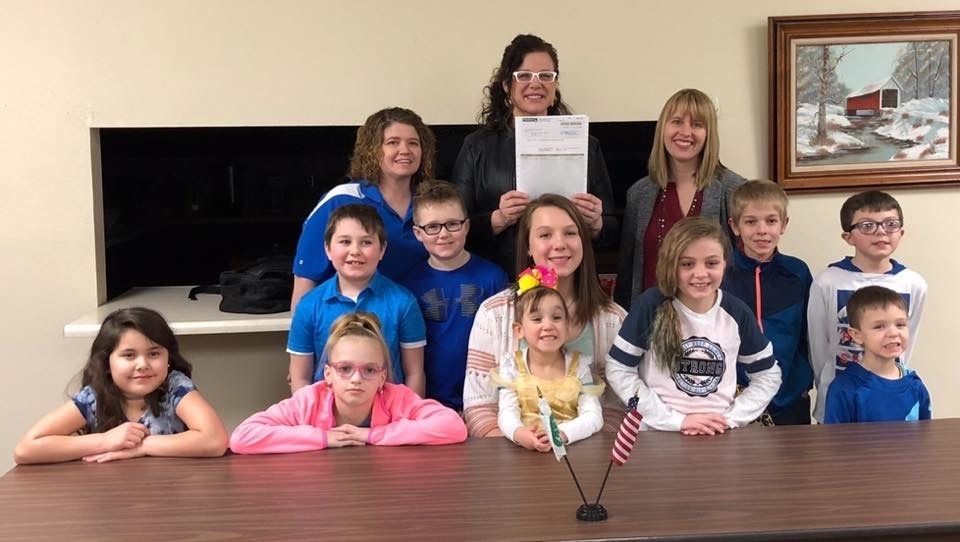 Group of children and adults pose behind a table, holding a large check.