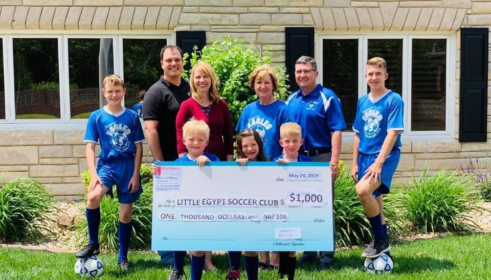 Group presents large check ($1,000) to youth soccer club. People in blue shirts, sunny outdoor setting.