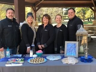 Five people stand behind a table with snacks and decorations outdoors. They wear black jackets.