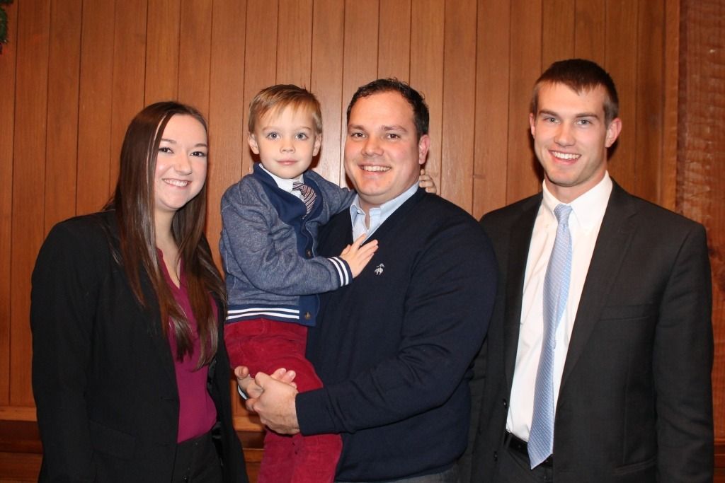 Four people smiling for a photo. A man holds a child; two people in suits stand beside them. Wooden background.