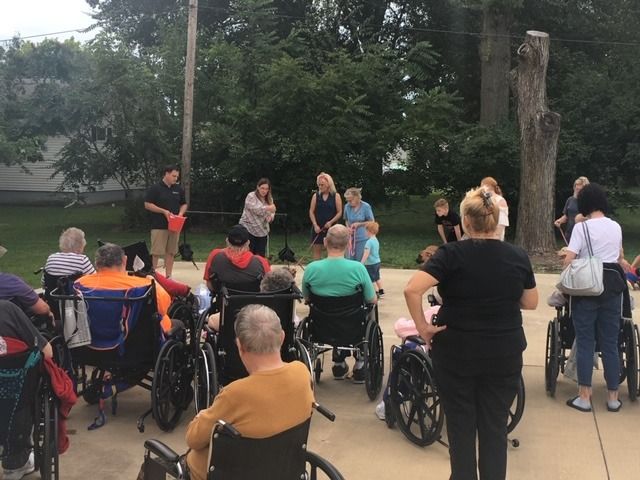People in wheelchairs and standing attend an outdoor event; trees and building in the background.