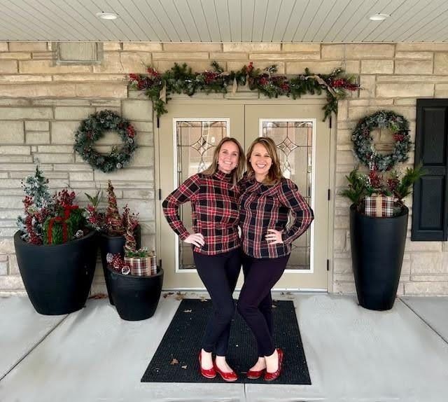 Two women in plaid shirts and black pants pose in front of a decorated doorway.