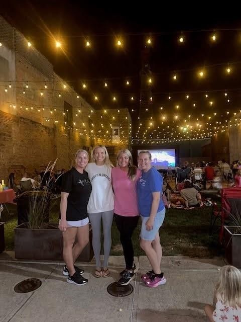 Four women pose under string lights at an outdoor movie night. People sit on blankets in the background.