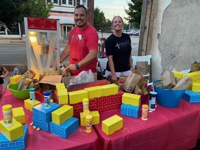 Two people selling popcorn at a street fair; red table, blue and yellow boxes, popcorn machine, and seasoning.