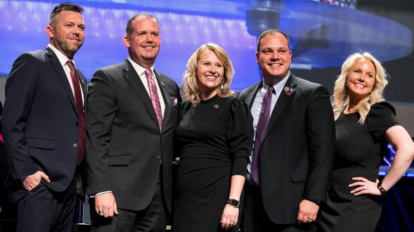 Five people smiling in formal attire, on a stage with a blurred background.