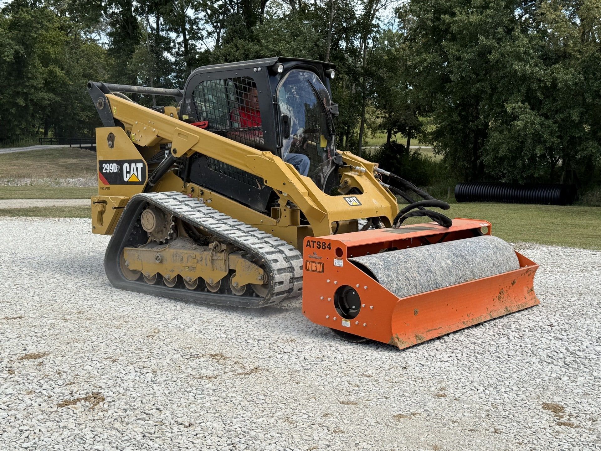 Yellow CAT skid steer with orange attachment parked on gravel in a wooded area