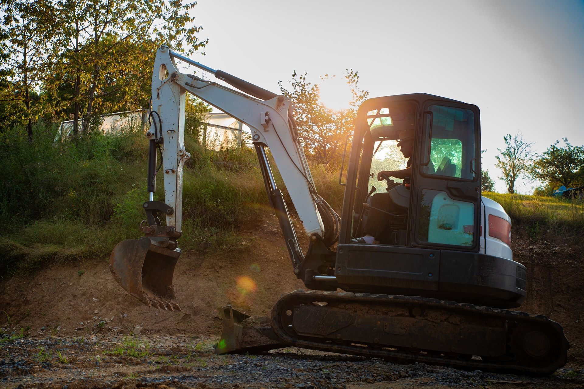 Excavator digging on a dirt path at sunset, with trees and bright sun in the background