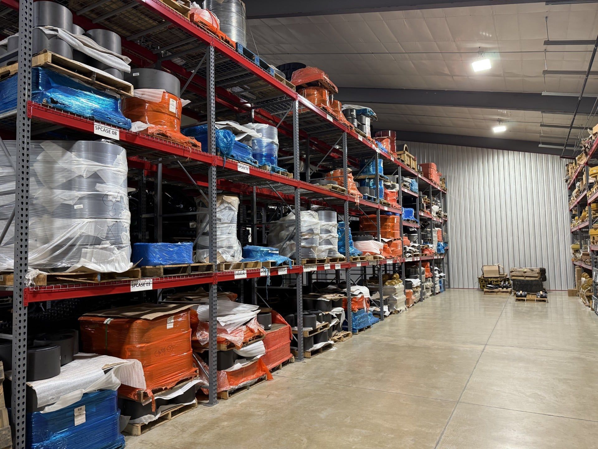 Warehouse aisle with tall metal racks of wrapped pallets and orange containers under bright lights.