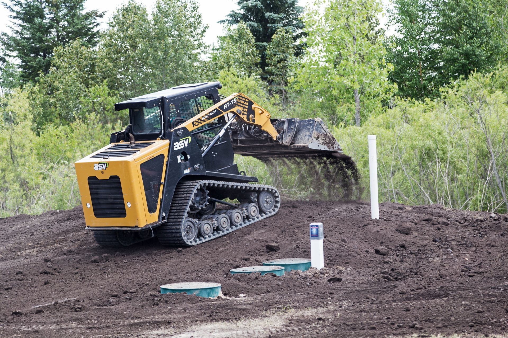 Yellow tracked skid steer leveling soil at a construction site near trees