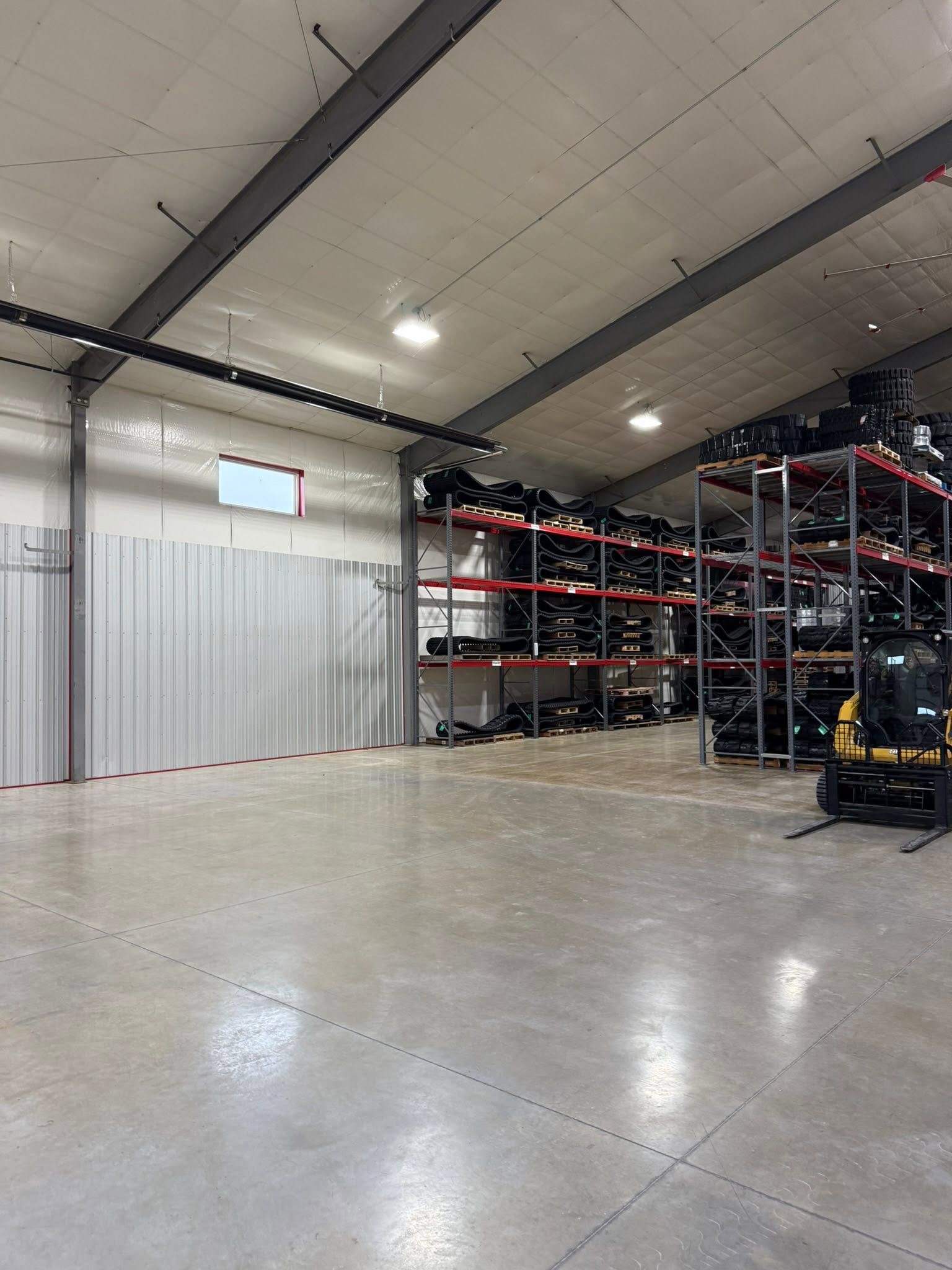 Warehouse aisle with tall metal racks of wrapped pallets and orange containers under bright lights.