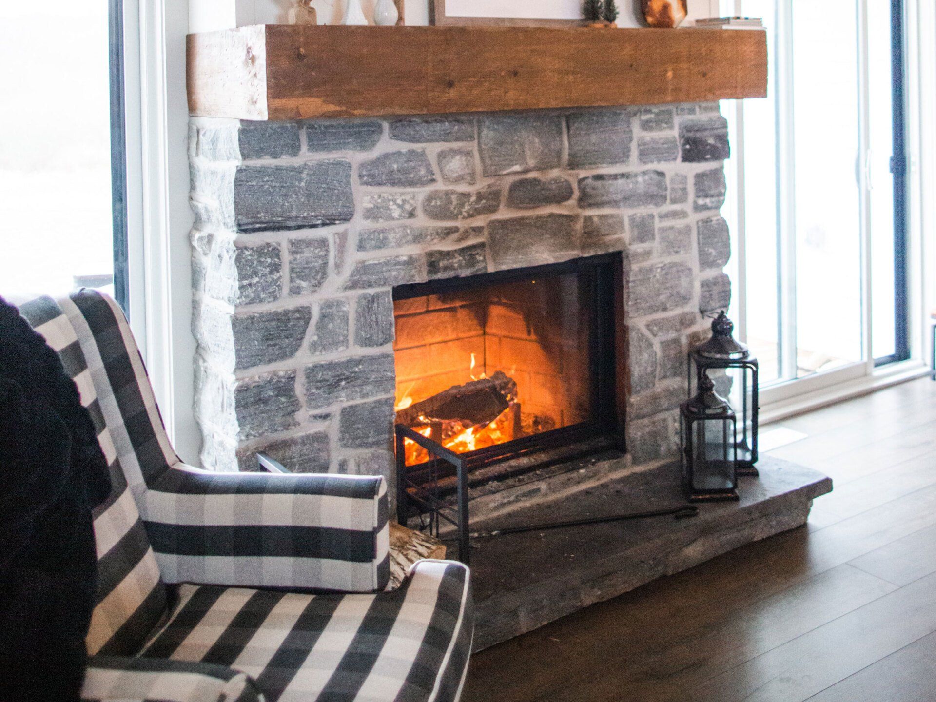Stone fireplace with a lit fire, wooden mantel, lantern, and black-and-white plaid chair in a living room.