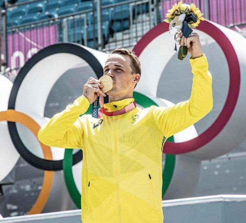 A man in a yellow jacket is holding a gold medal in front of the olympic rings