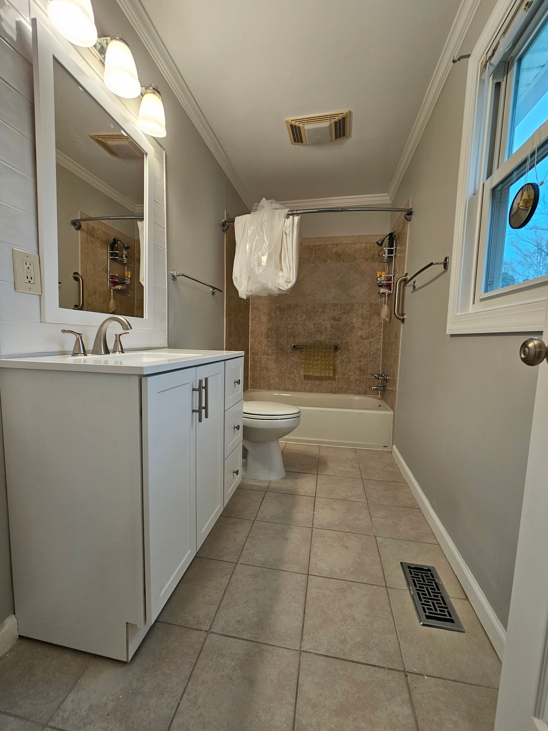 Narrow bathroom with vanity, toilet, bathtub, tan tile, and a window letting in daylight.