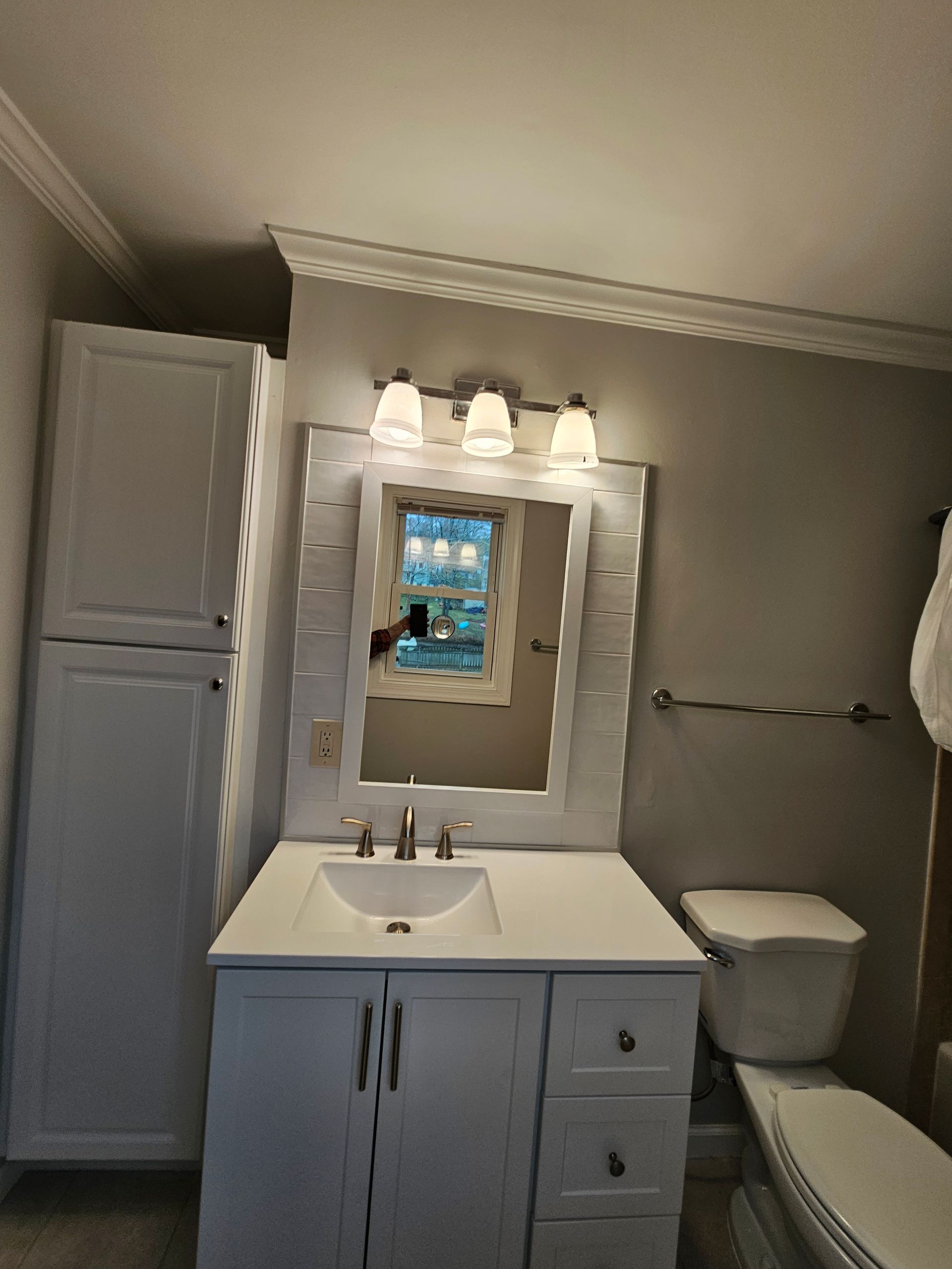 Small bathroom with white vanity, mirror lights, toilet, towel bar, and a window reflected in the mirror.