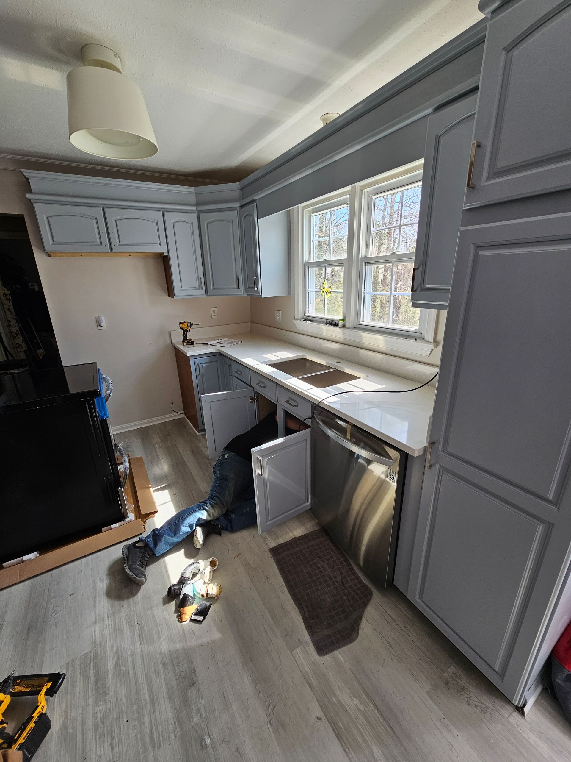 Narrow kitchen with gray cabinets, white countertops, and a window over the sink, with tools and items on the floor.