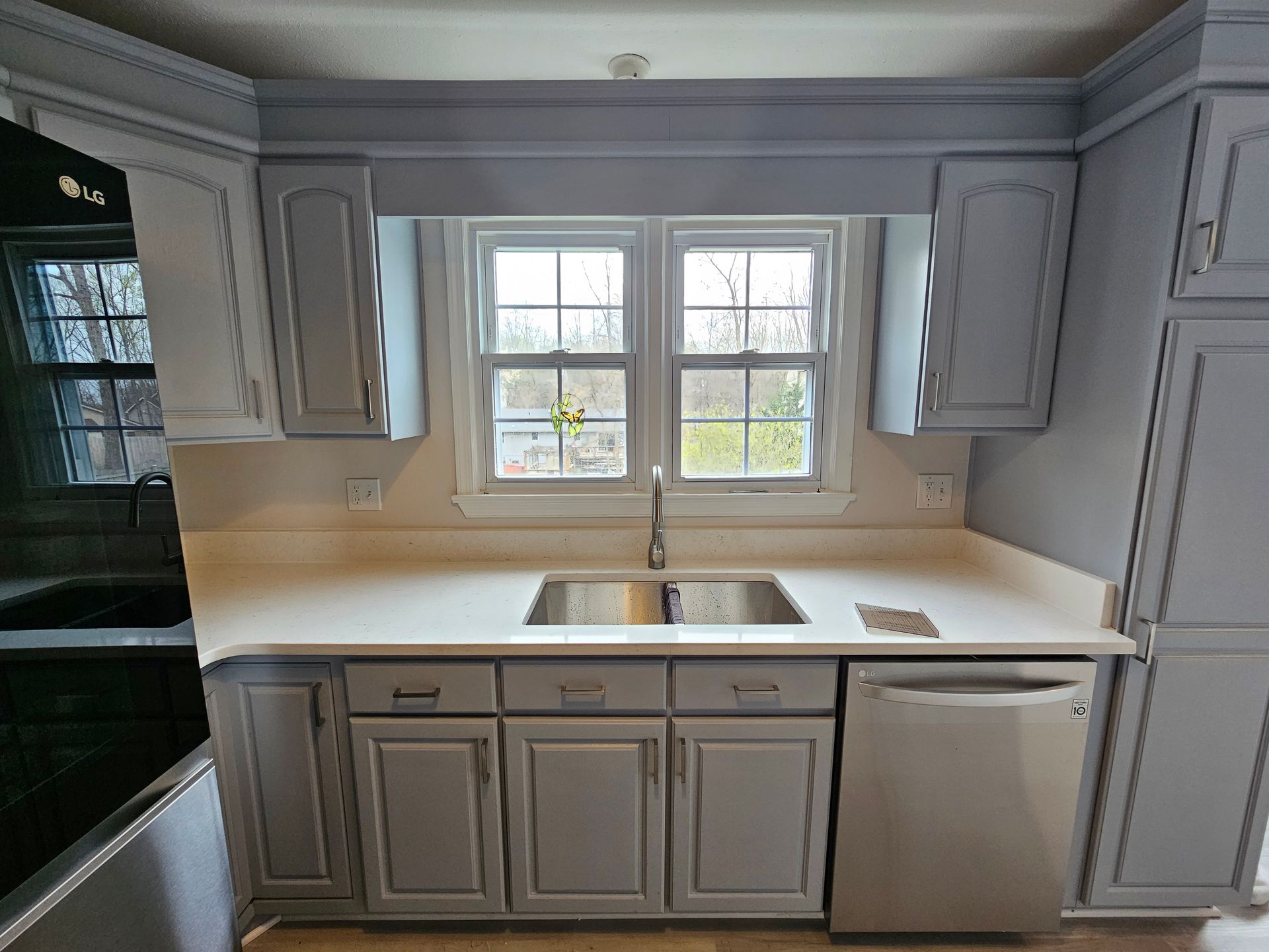 Bright kitchen with gray cabinets, white countertops, double window, sink, and stainless dishwasher.