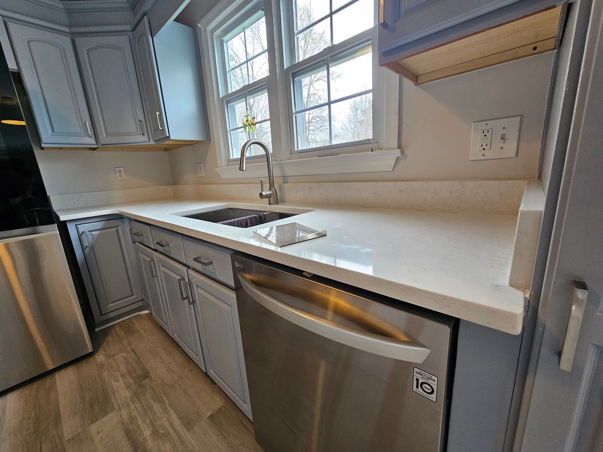 Bright kitchen with white countertops, gray cabinets, stainless sink, and wood floor by a window