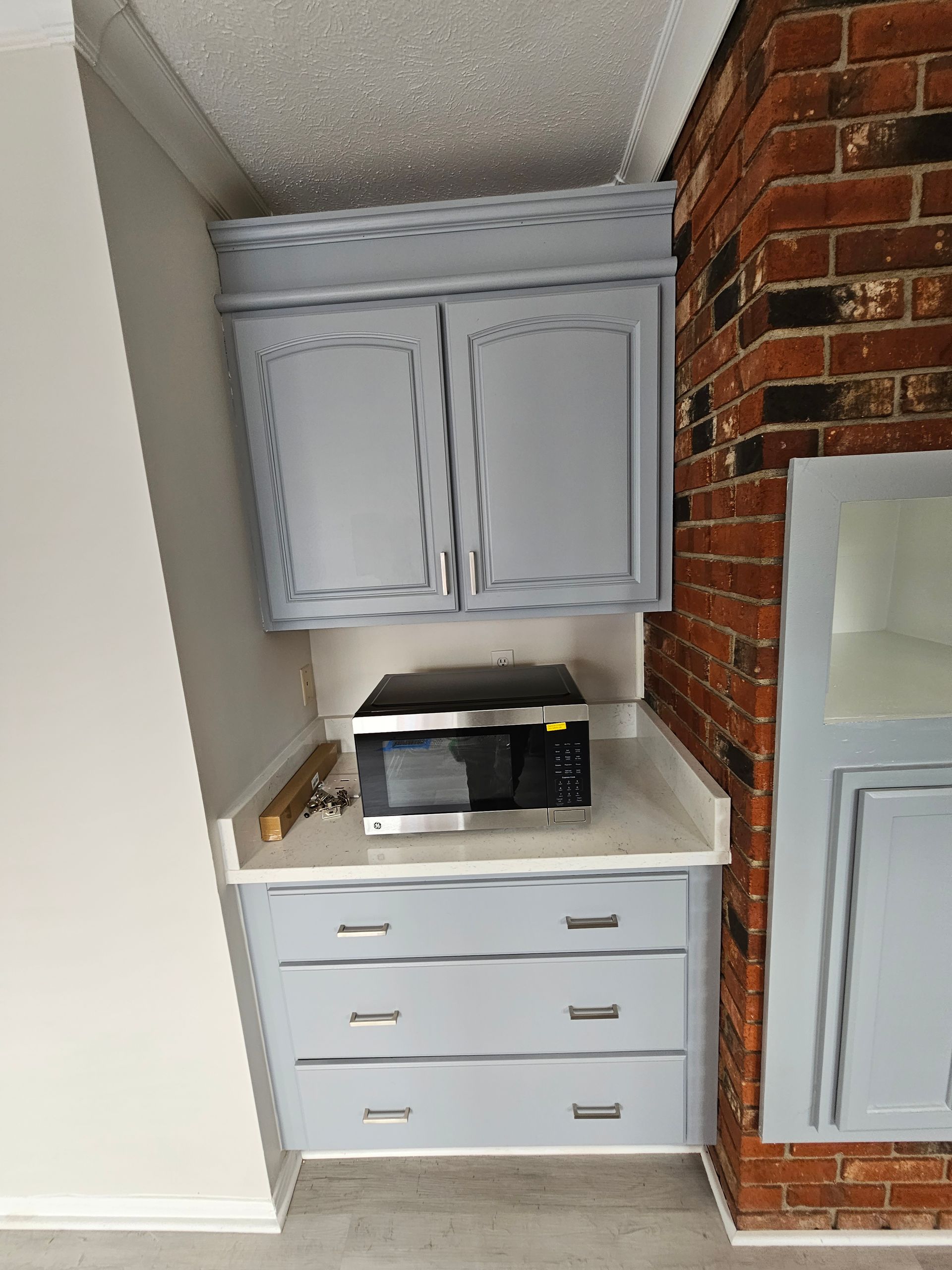 Small kitchen nook with gray cabinets, white countertop, microwave, and exposed brick wall.