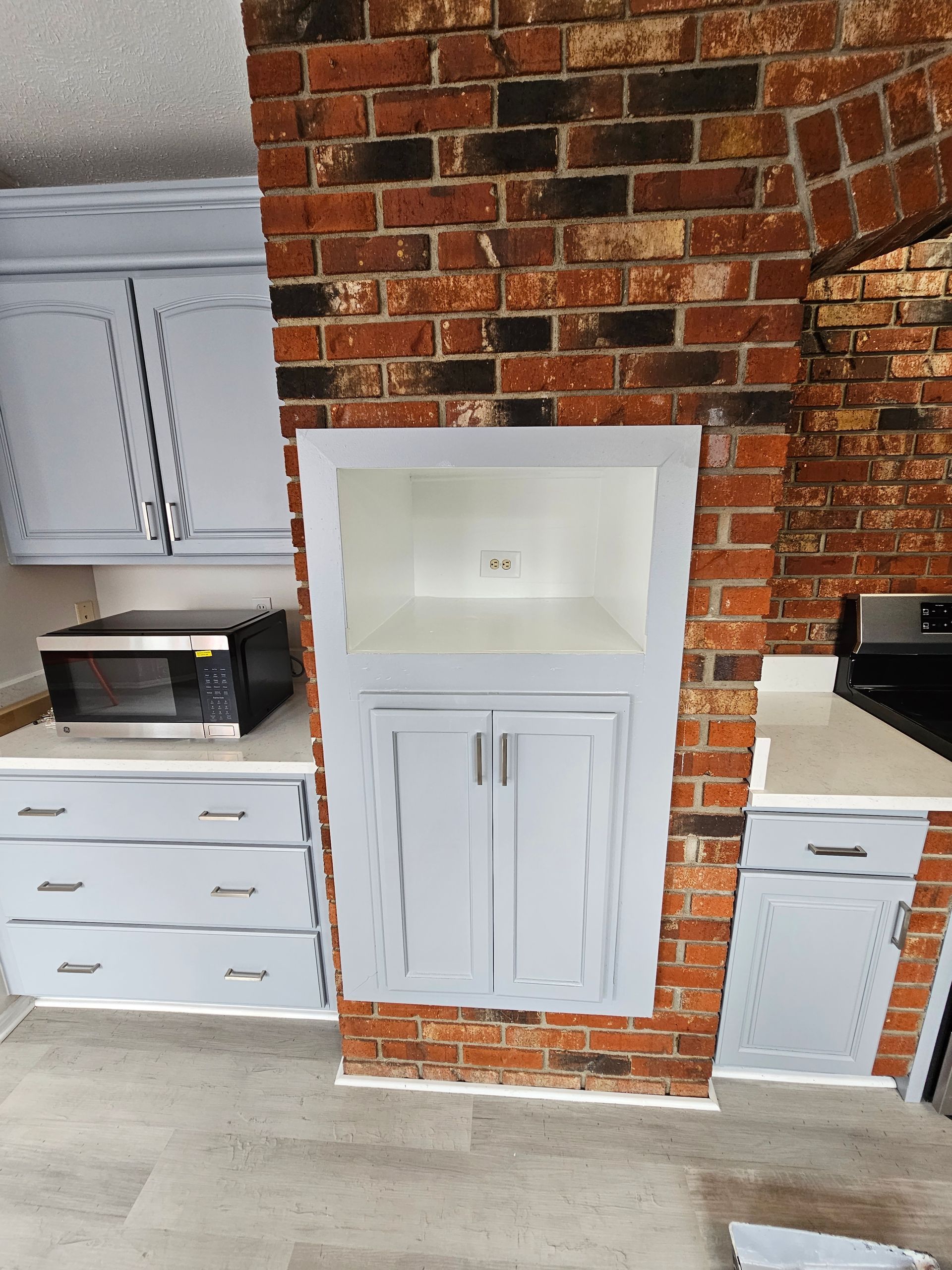 Built-in white cabinet niche in a brick wall, flanked by light-colored kitchen cabinets.