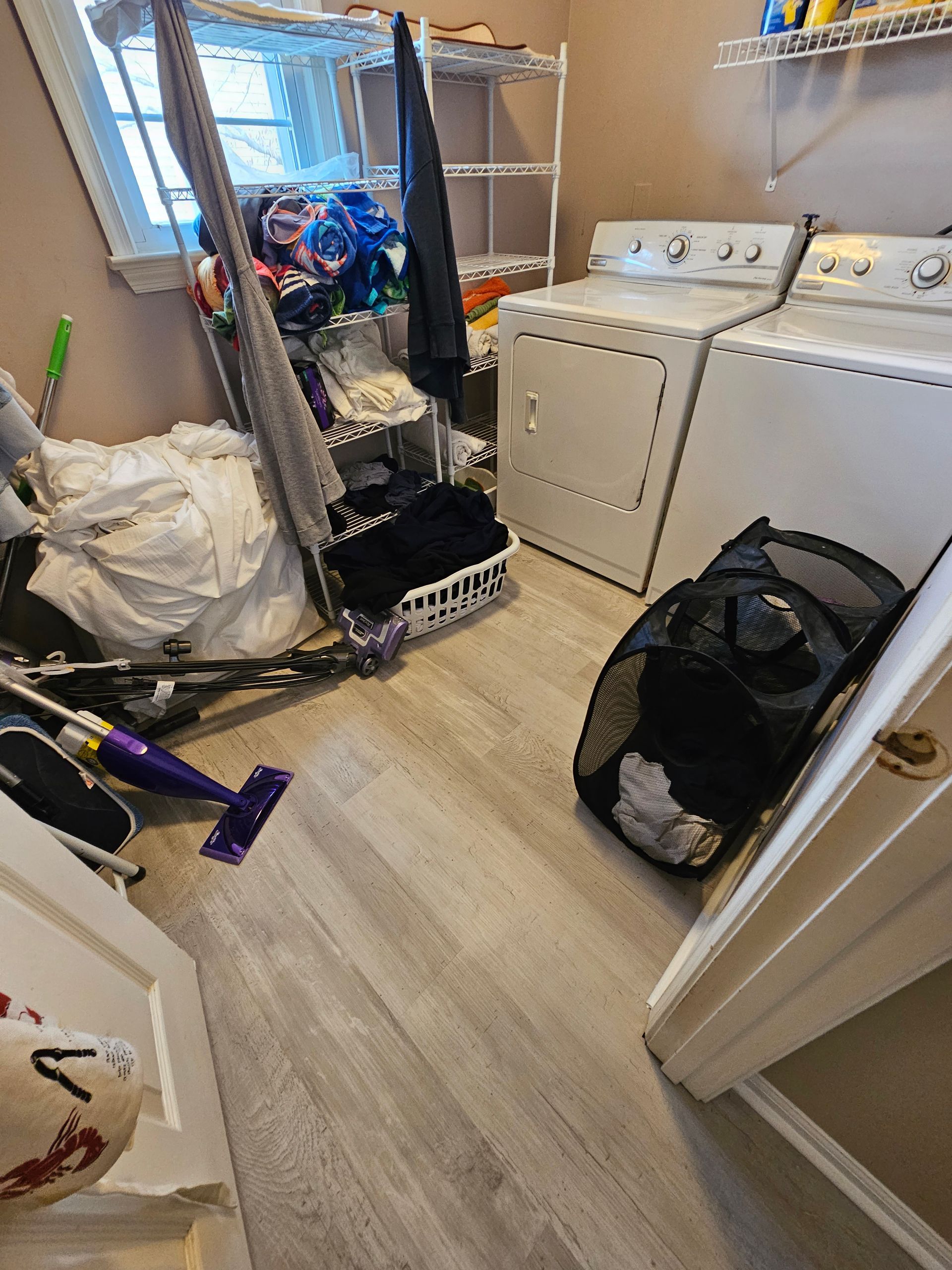 Laundry room with washer and dryer, shelf, cleaning supplies, and cluttered floor with bags and tools.