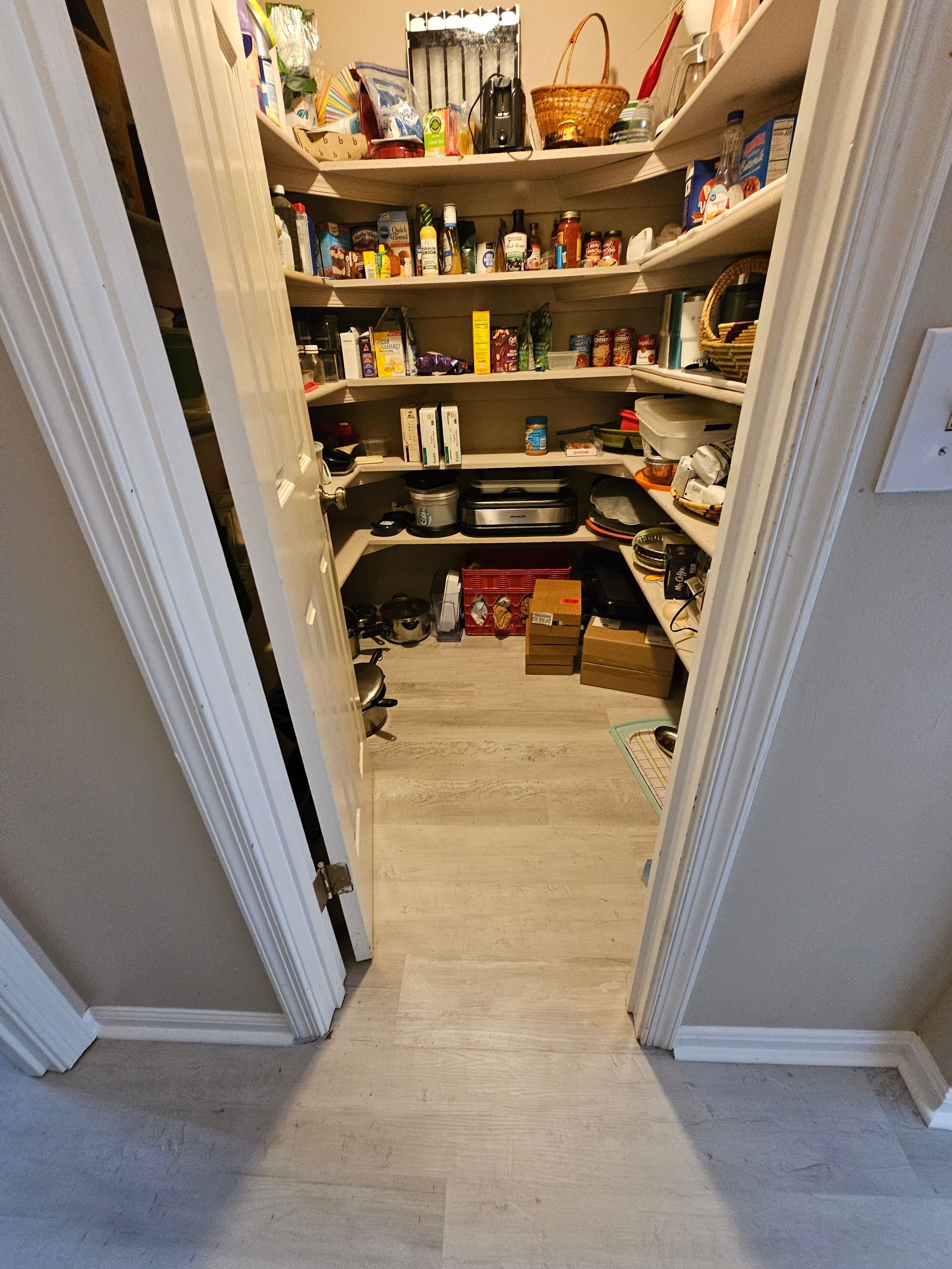 Narrow pantry with shelves packed with food, bottles, and kitchen items, viewed through open doorway.