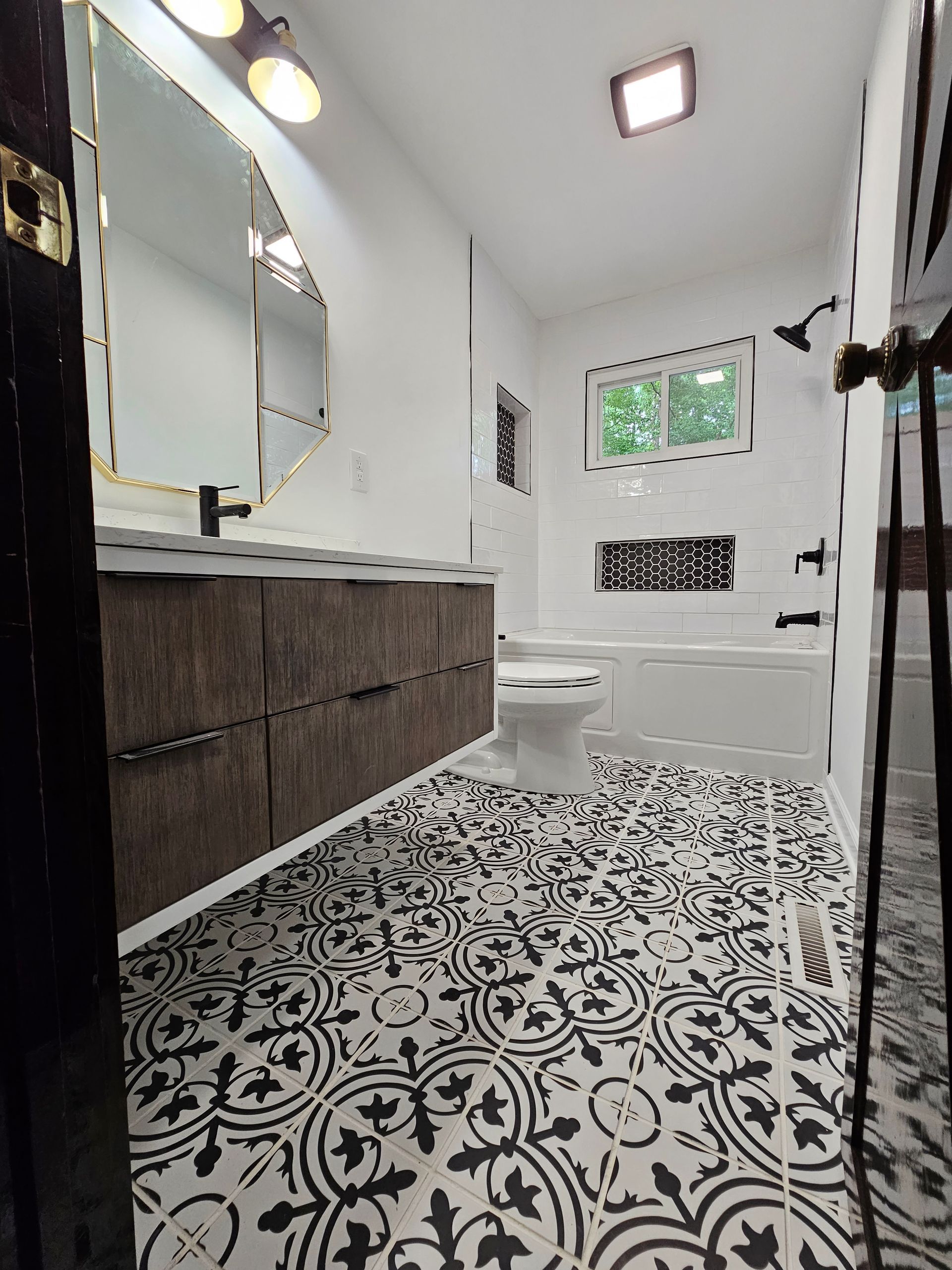 Modern bathroom with patterned black-and-white tile, vanity mirror, toilet, and window.