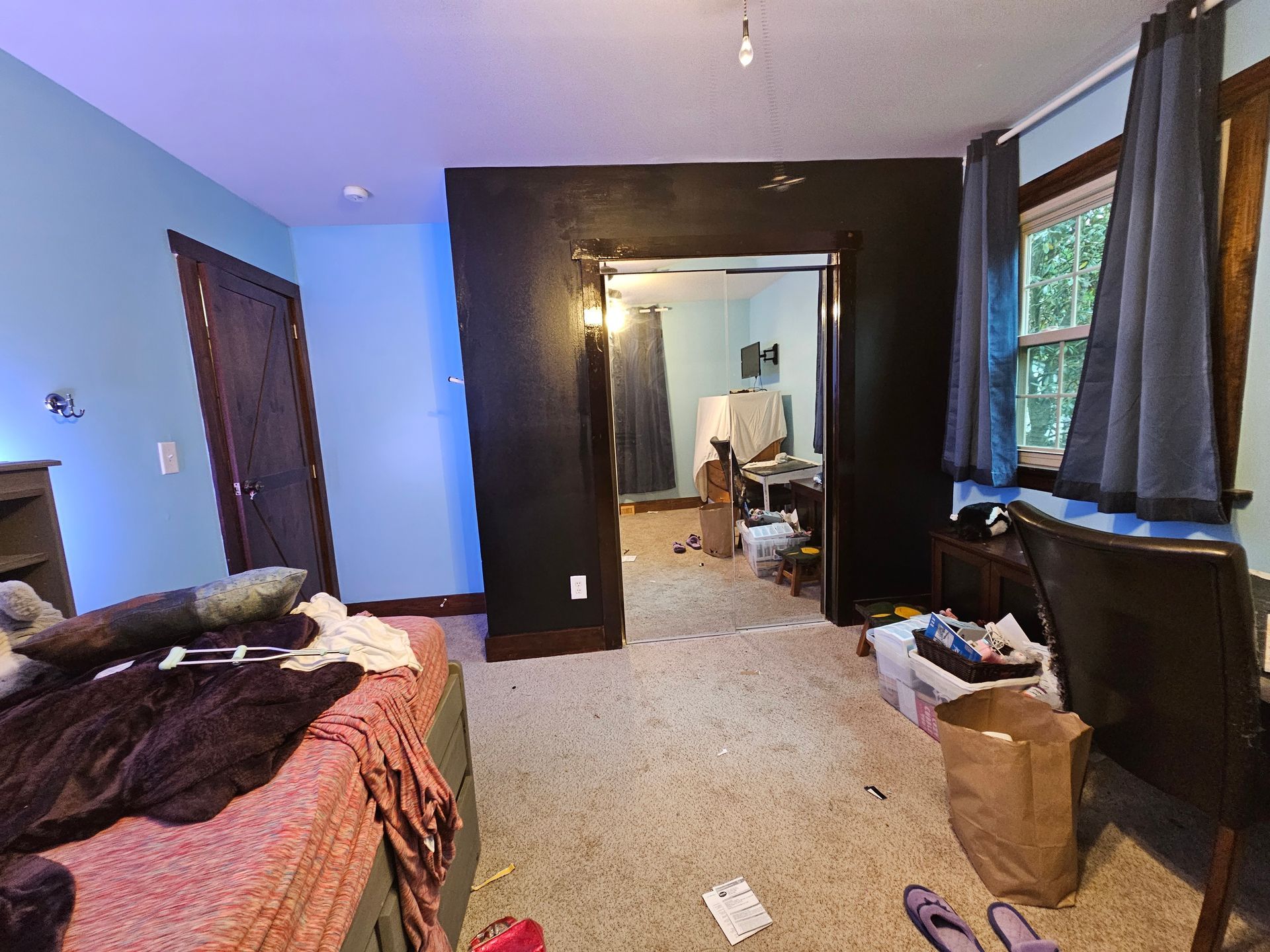 Messy bedroom with bed, carpet, mirror, chair, and boxes near a window and doorway.