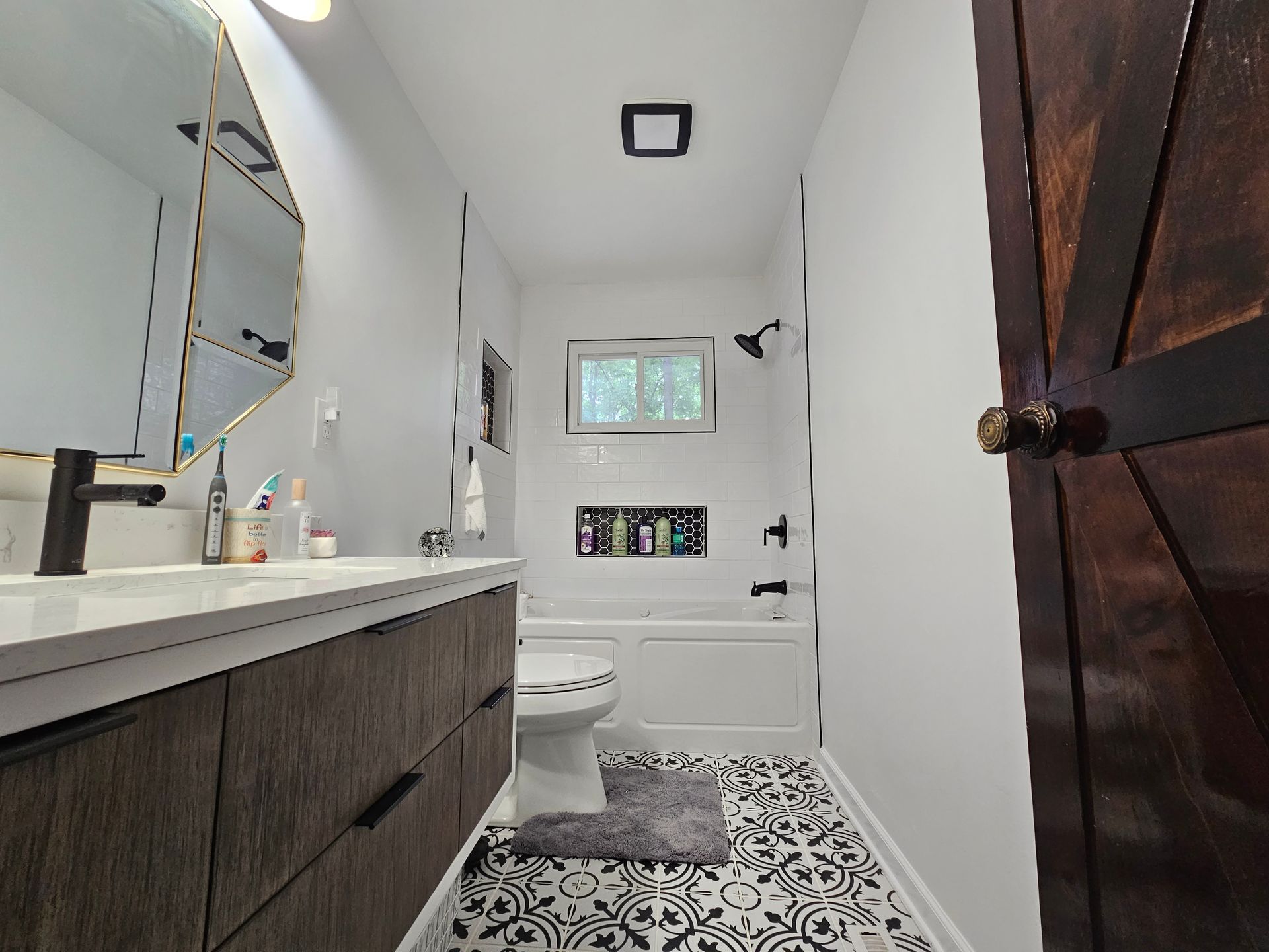 Narrow modern bathroom with white walls, dark vanity, patterned floor, toilet, shower, and window.