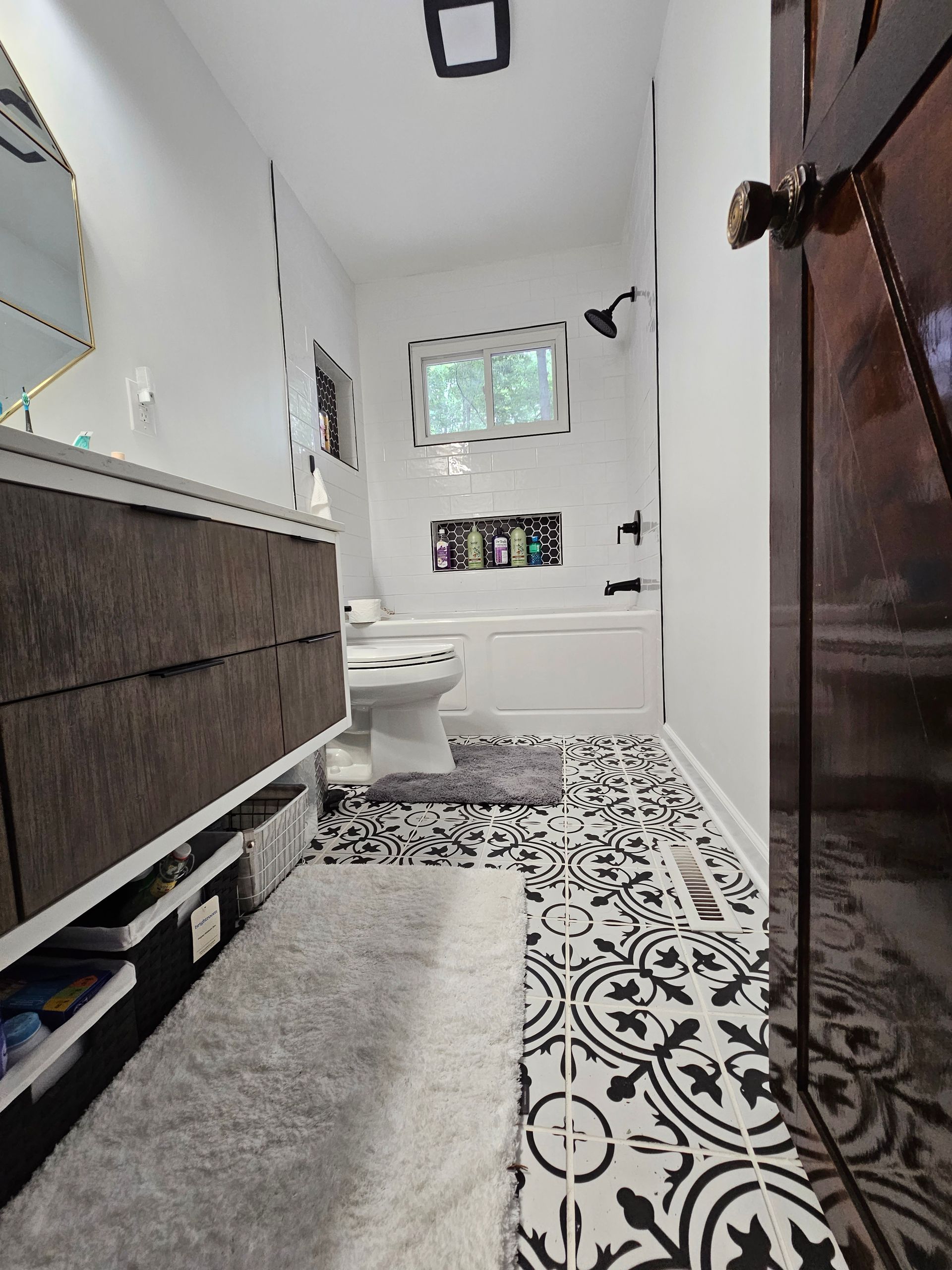 Narrow bathroom with patterned tile floor, vanity, toilet, and window above the tub.