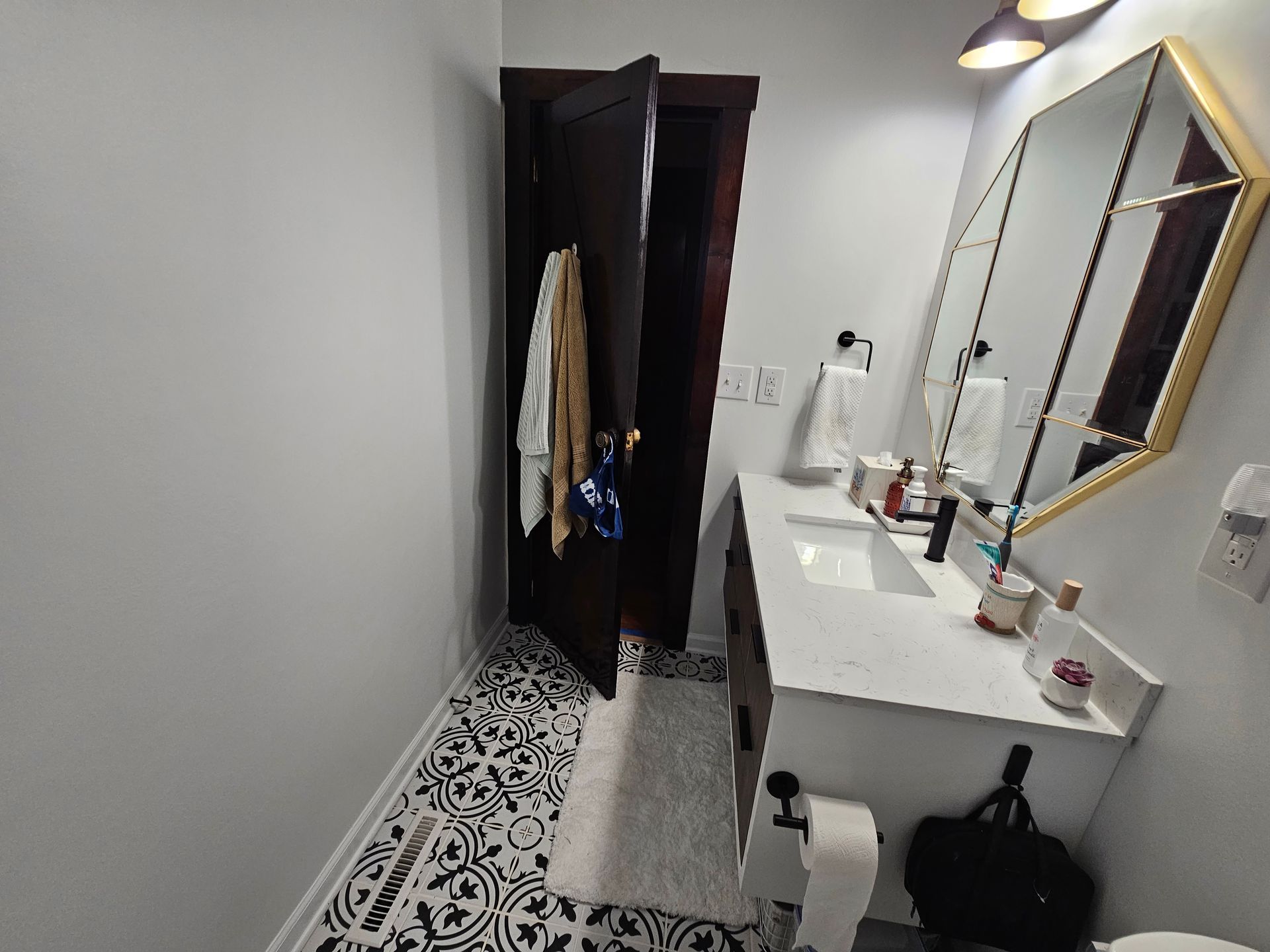 Narrow bathroom with black-and-white patterned floor, white vanity, mirror, and open door to a hanging closet