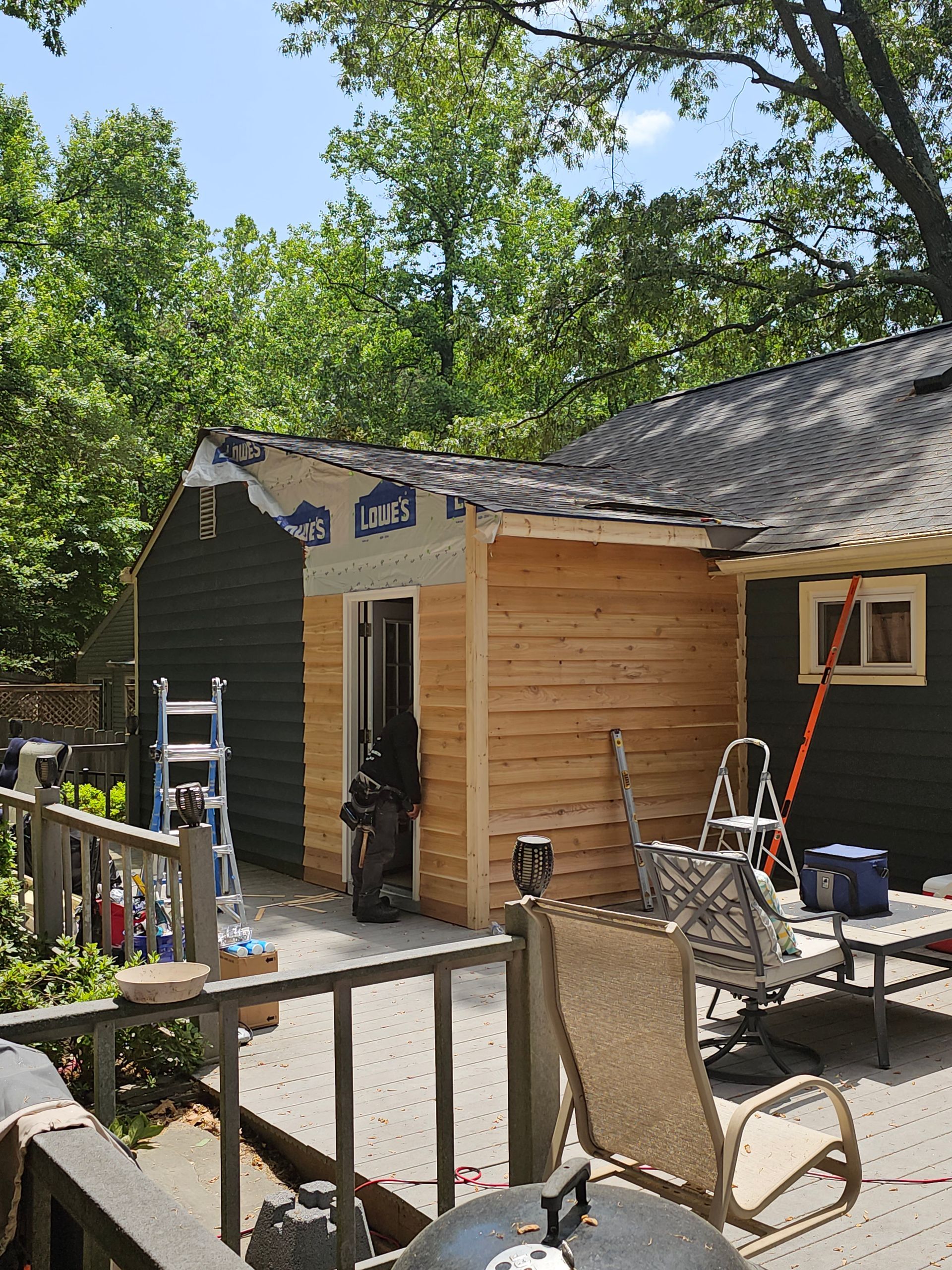 Backyard patio with a small shed, ladder, chairs, and tools beside a wooden fence and trees