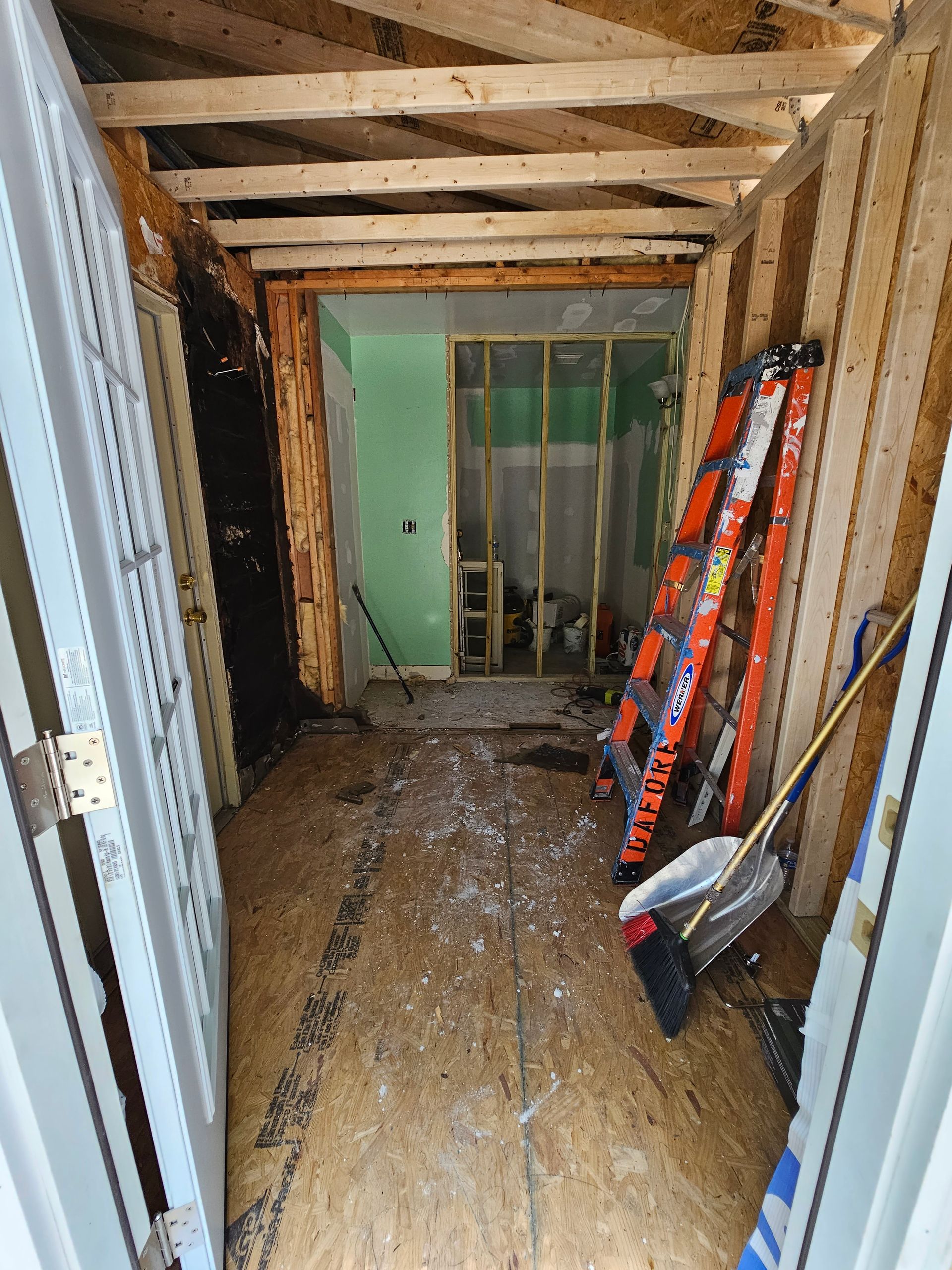 Narrow unfinished hallway with exposed wood beams, corrugated metal walls, and tools leaning against the right wall.