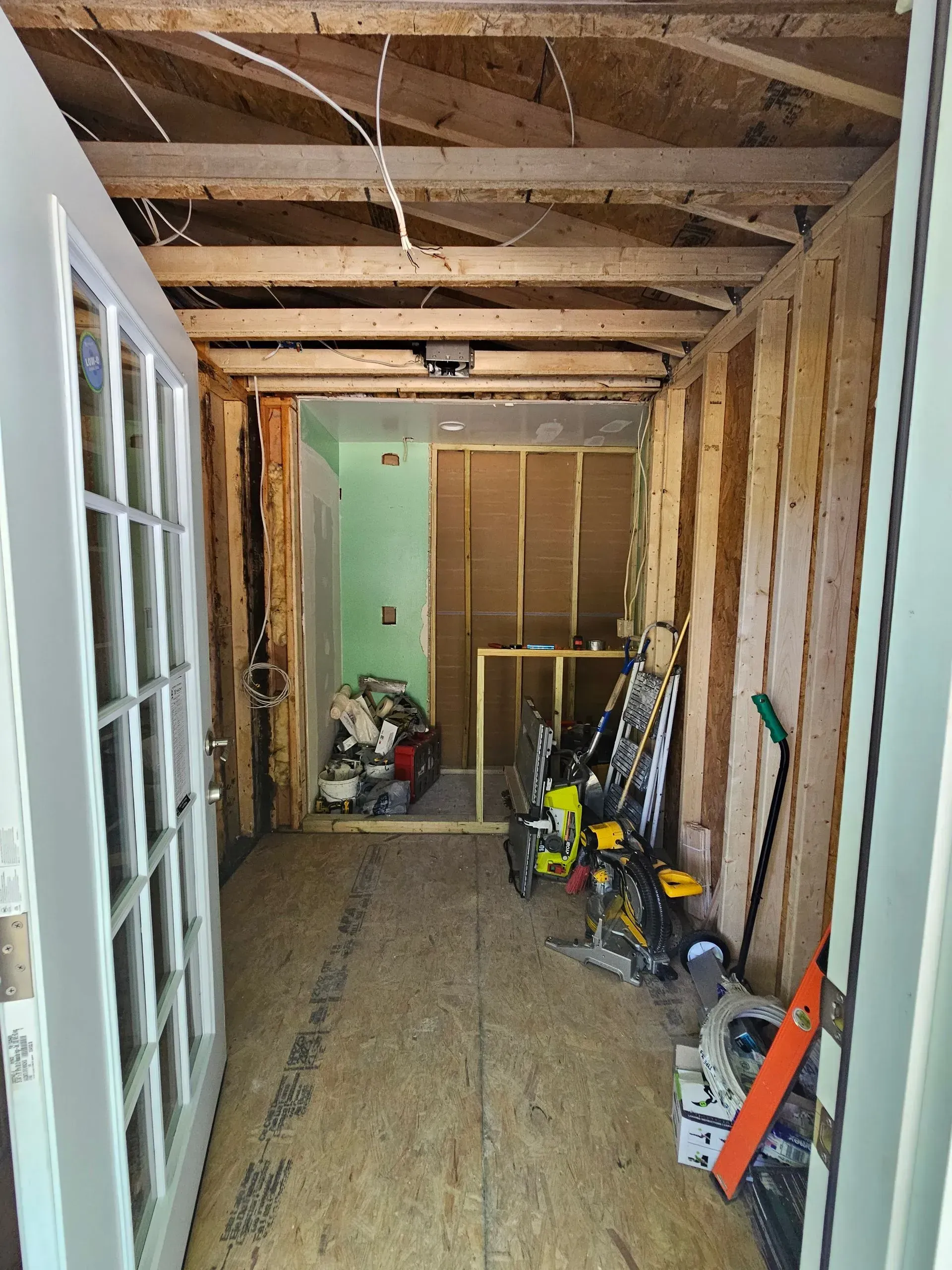 Narrow unfinished hallway with exposed studs, tools, and debris on the floor leading to a green door.