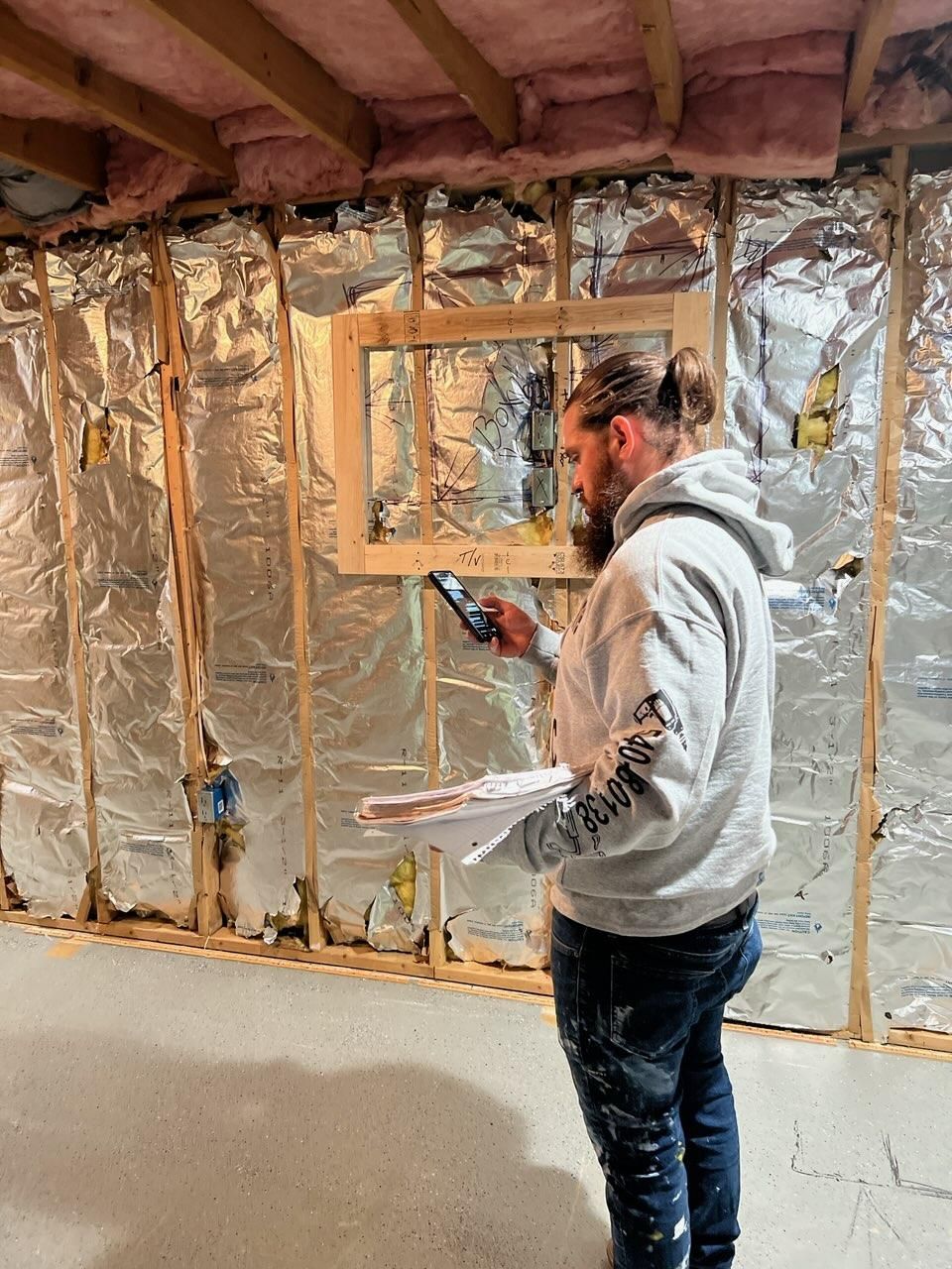 Person in a basement under renovation holding papers, standing beside exposed insulation and a framed window opening.