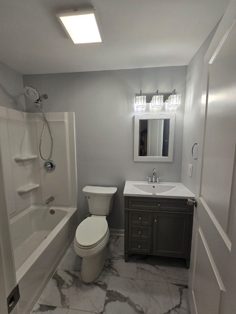 Small gray bathroom with a tub, toilet, vanity, and mirror under bright ceiling light.