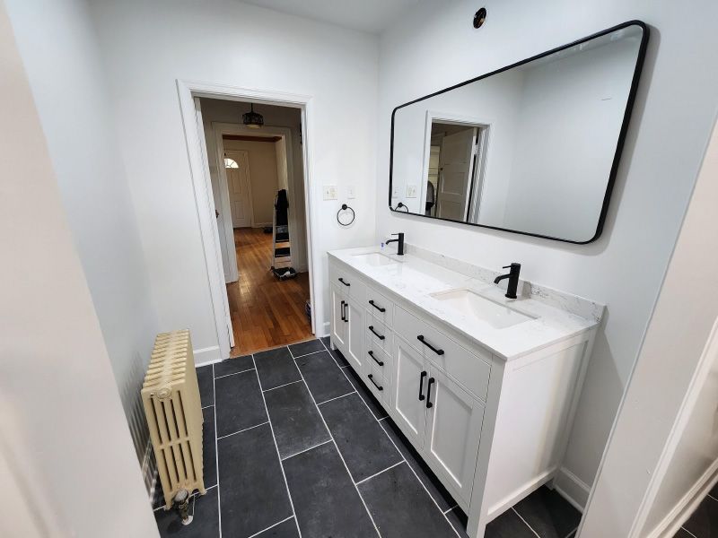 Modern bathroom with white vanity, large mirror, black tile floor, and hallway visible through open door