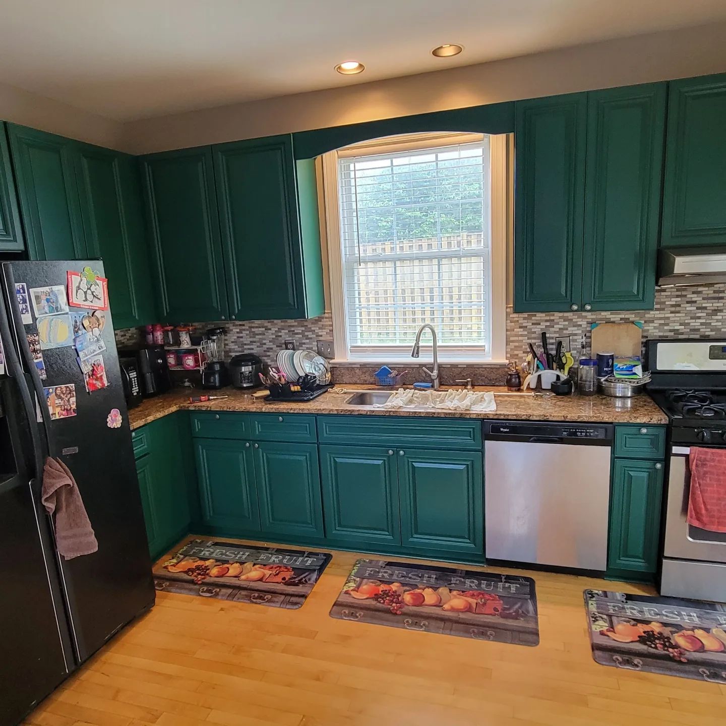 Kitchen with teal cabinets, black fridge, sink under window, and colorful rugs on wood floor.