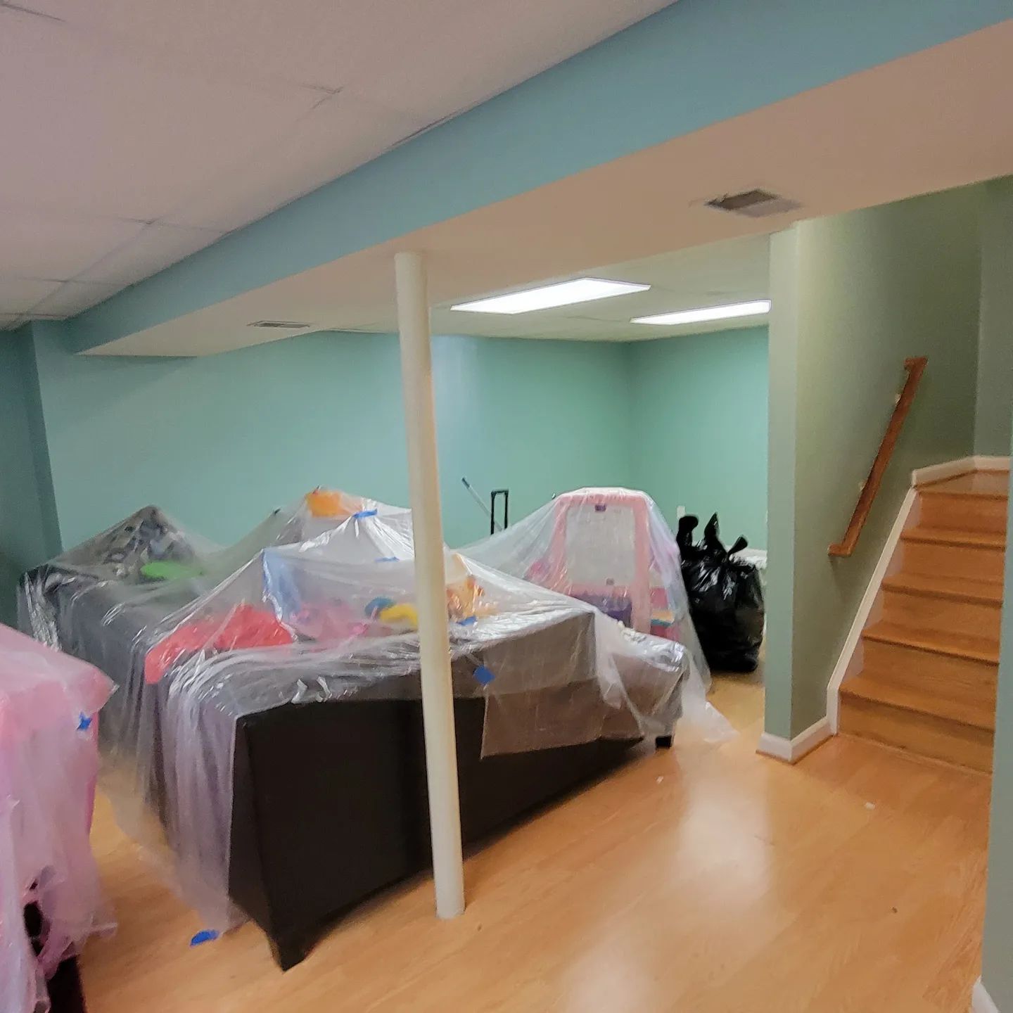 Basement room with covered furniture, teal walls, wood floor, and stairs on the right.