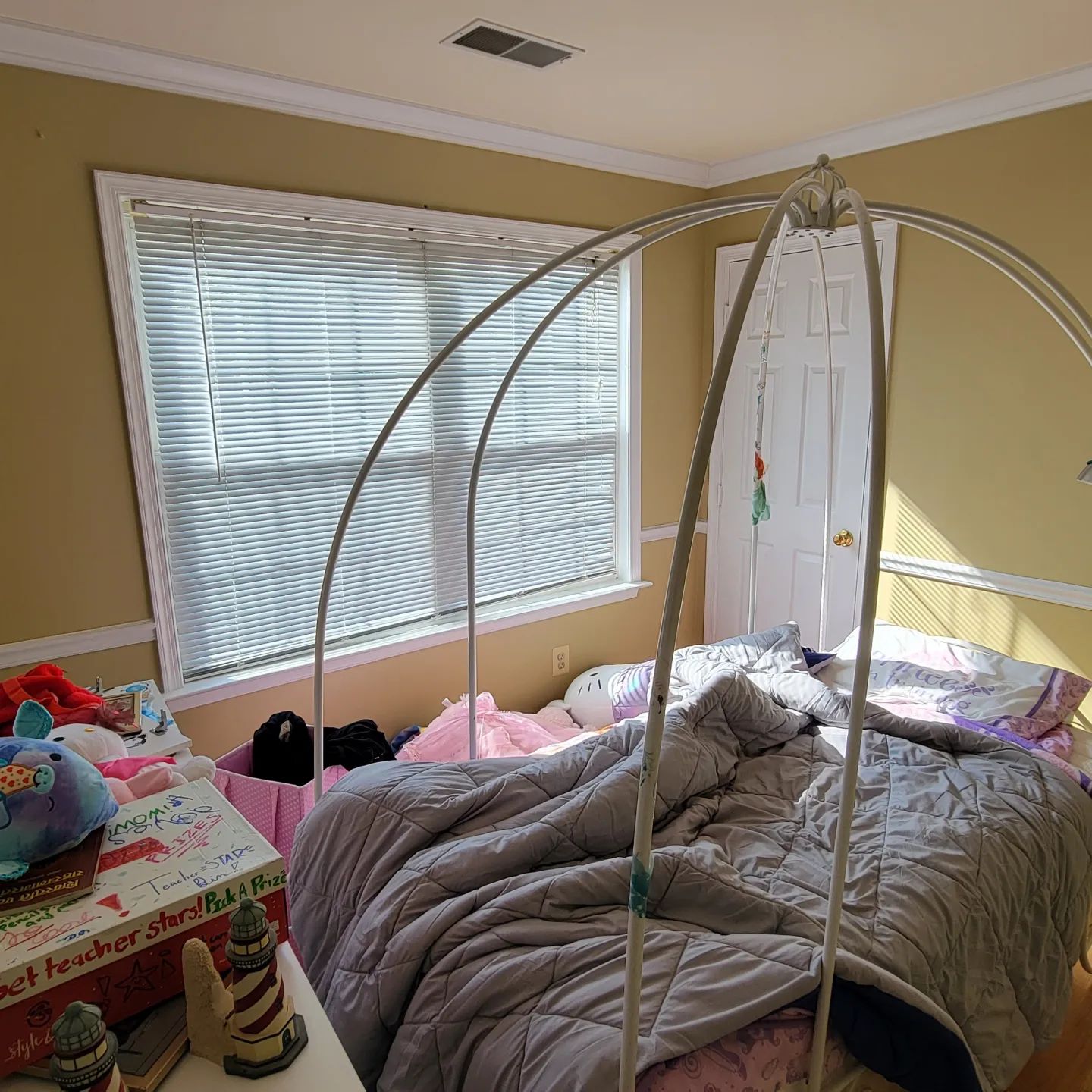 Bedroom with unmade bed, white canopy frame, gray comforter, and window blinds on tan walls.